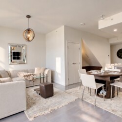 Open-plan living and dining area with a beige sectional sofa, glass coffee table, and a round dining table with white chairs near a kitchen island.