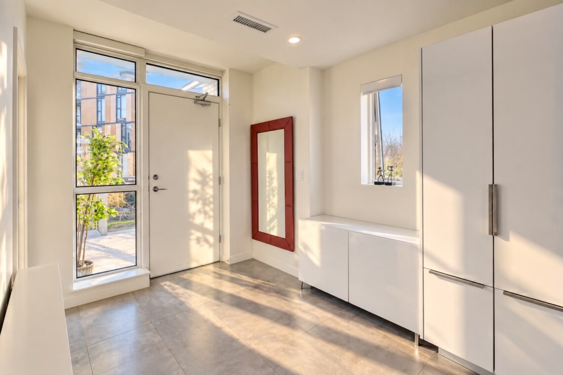 Bright, modern entryway with white cabinets, a pale front door, and a red-framed wall mirror beside a small window. Outside visible plants and building panels.