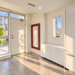 Bright, modern entryway with white cabinets, a pale front door, and a red-framed wall mirror beside a small window. Outside visible plants and building panels.