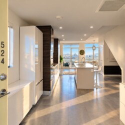 Open-concept kitchen with white cabinets, an island, and bar stools, leading to large windows overlooking water and city skyline.