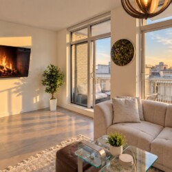 Sunlit modern living room with a wall-mounted fireplace, beige sofa, and large windows opening to a balcony at sunset.
