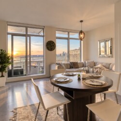 Open-plan dining and living area with a round table set for four, beige sofa, and sunset balcony view.
