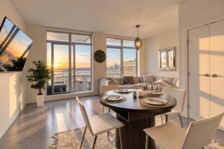 Open-plan dining and living area with a round table set for four, beige sofa, and sunset balcony view.