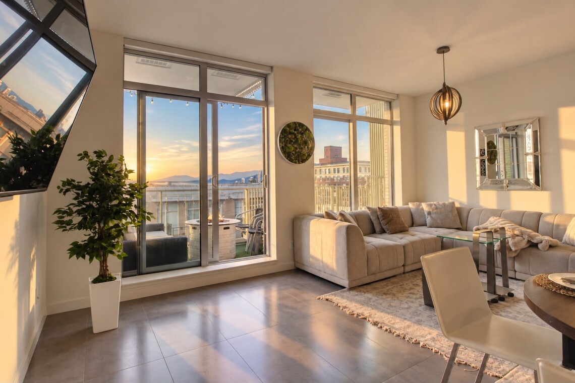 Sunset-lit living room with a beige L-shaped sofa, glass coffee table, and floor-to-ceiling windows opening to a balcony view of buildings and mountains.