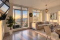 Sunset-lit living room with a beige L-shaped sofa, glass coffee table, and floor-to-ceiling windows opening to a balcony view of buildings and mountains.