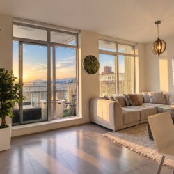 Sunset-lit living room with a beige L-shaped sofa, glass coffee table, and floor-to-ceiling windows opening to a balcony view of buildings and mountains.