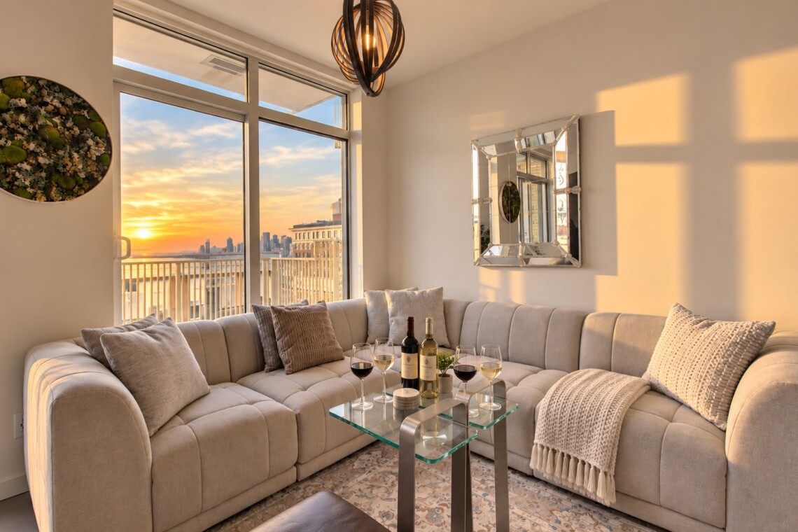 Sunlit modern living room with a beige sectional, glass coffee table, and sunset view of a city skyline through large windows.