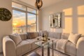 Sunlit modern living room with a beige sectional, glass coffee table, and sunset view of a city skyline through large windows.