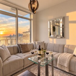 Sunlit modern living room with a beige sectional, glass coffee table, and sunset view of a city skyline through large windows.