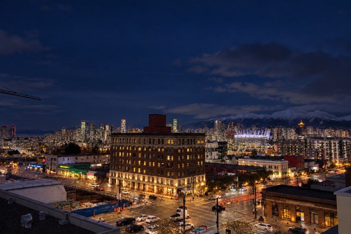 Night cityscape at dusk with illuminated streets, a cluster of tall buildings, a bright arena, and snow-capped mountains in the distance.