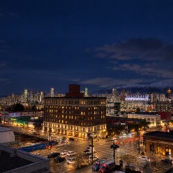 Night cityscape at dusk with illuminated streets, a cluster of tall buildings, a bright arena, and snow-capped mountains in the distance.