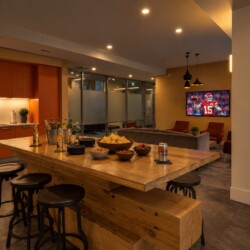 Modern kitchen and lounge with a long wooden island, chip bowls, drinks, and a wall-mounted TV showing a football game.