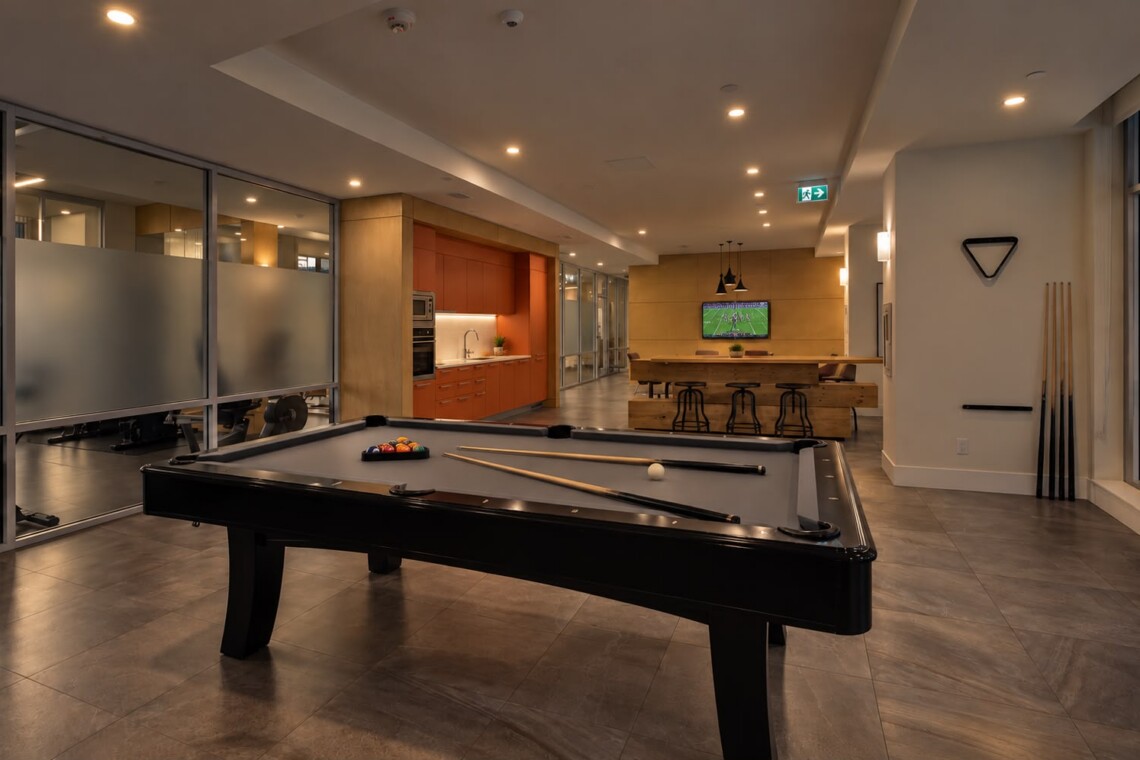 Open, modern recreation area with a black pool table in the foreground, a warm kitchenette to the left, and a TV mounted on a wooden wall in the background.