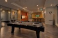 Open, modern recreation area with a black pool table in the foreground, a warm kitchenette to the left, and a TV mounted on a wooden wall in the background.