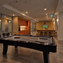Open, modern recreation area with a black pool table in the foreground, a warm kitchenette to the left, and a TV mounted on a wooden wall in the background.