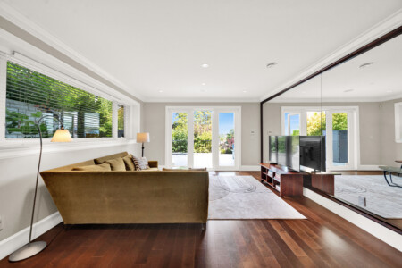 Bright, modern living room with a brown sofa facing a wall of mirrored panels and a TV stand; large windows show greenery outside.
