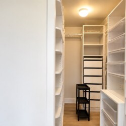 Bright walk-in closet with white built-in shelves, drawers, and a small black two-tier stool in the center of the room area.