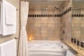 Tub-and-shower combo in warm beige tile bathroom with a candle on the tub edge and white towels on a wall rack in a spa-like setting.
