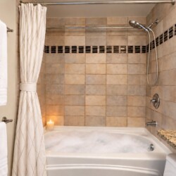 Tub-and-shower combo in warm beige tile bathroom with a candle on the tub edge and white towels on a wall rack in a spa-like setting.