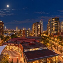 Nighttime cityscape with lit high-rise buildings along a street, a plaza with a white sail-shaped canopy in the foreground, and a bright full moon in a dark blue sky.
