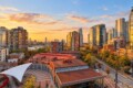 Sunset over a modern city skyline with tall glass towers, a red-roofed building, and a white tensile canopy in a plaza foreground.