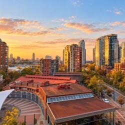 Sunset over a modern city skyline with tall glass towers, a red-roofed building, and a white tensile canopy in a plaza foreground.