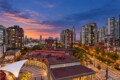 Sunset over a modern cityscape with high-rise residential towers along a waterfront and a curved plaza with a white canopy in the foreground.
