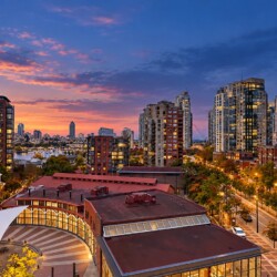 Sunset over a modern cityscape with high-rise residential towers along a waterfront and a curved plaza with a white canopy in the foreground.