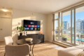 Bright living room with wall-mounted Apple TV, round black table, two beige chairs, and a plant, plus sliding glass doors to a balcony with a city skyline view.