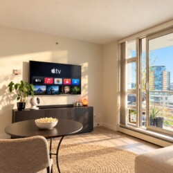 Bright living room with wall-mounted Apple TV, round black table, two beige chairs, and a plant, plus sliding glass doors to a balcony with a city skyline view.