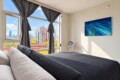 Bright modern bedroom with a large window view of a city skyline, gray drapes, and a bed with white pillows and a dark gray comforter, plus a blue abstract painting on the wall.