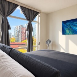 Bright modern bedroom with a large window view of a city skyline, gray drapes, and a bed with white pillows and a dark gray comforter, plus a blue abstract painting on the wall.