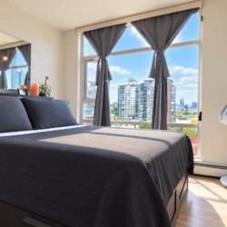 Modern bedroom with a dark gray bed and large window showing the city skyline; gray curtains pulled back.
