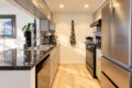 Modern galley kitchen with stainless steel fridge, oven, and dishwasher, light wood flooring, and a marble-like countertop. A colorful quilted apron hangs on the back wall beside a small plant and framed seaside artwork.