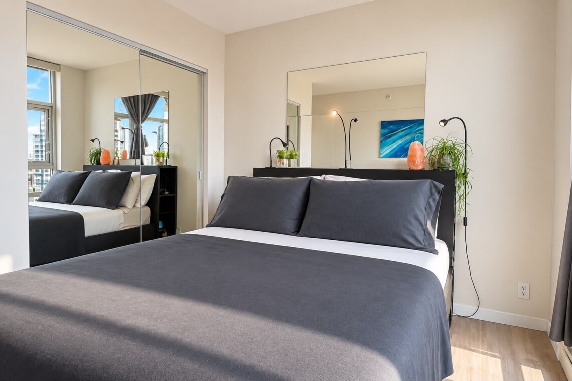 Modern bedroom with a gray-ink bed, mirrored wardrobe, and large window showing city buildings outside of sheer curtains.