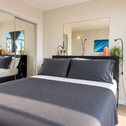 Modern bedroom with a gray-ink bed, mirrored wardrobe, and large window showing city buildings outside of sheer curtains.