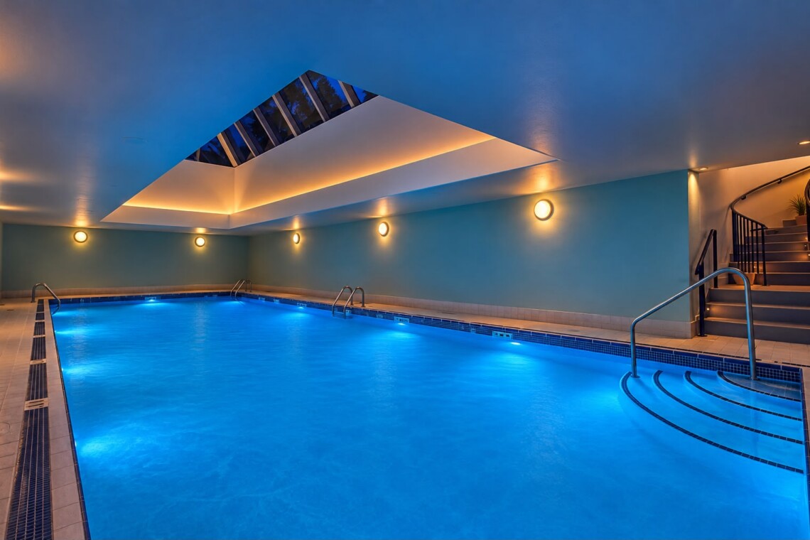 Indoor swimming pool with blue-lit water, beige tiled surround, and a curved staircase at the right edge.