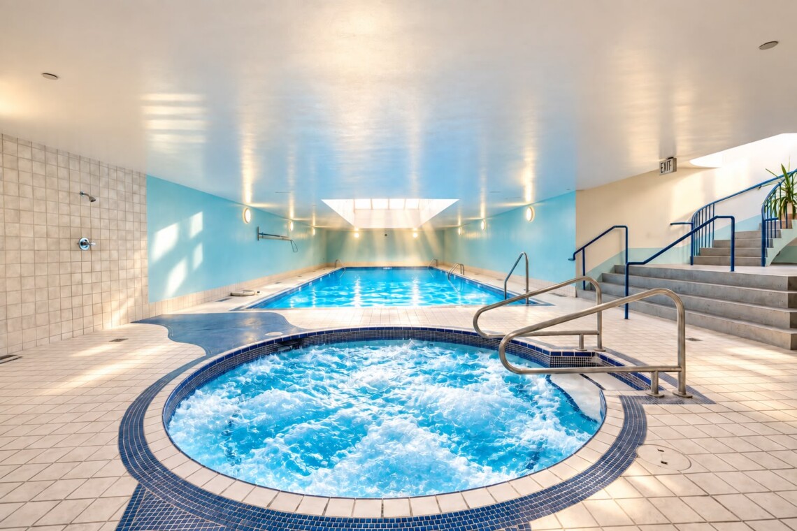 Indoor spa area with a circular hot tub in the foreground and a rectangular pool beyond, blue walls, metal railings, and stairs to the right.