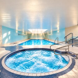 Indoor spa area with a circular hot tub in the foreground and a rectangular pool beyond, blue walls, metal railings, and stairs to the right.