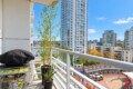 Balcony with a covered grill on a cart and potted plants, overlooking a city skyline of tall glass buildings and a marina on a sunny day.