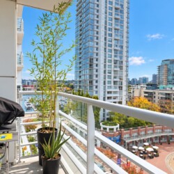 Balcony with a covered grill on a cart and potted plants, overlooking a city skyline of tall glass buildings and a marina on a sunny day.