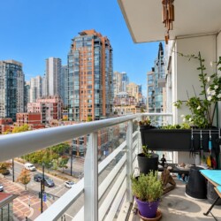 Balcony with blue lounge chair, potted plants, and urban city skyline of tall glass buildings across the street on a sunny day