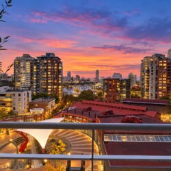 City skyline at sunset with orange-pink sky, modern high-rise buildings, and a railing in the foreground.