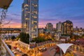 Urban waterfront at dusk with tall illuminated buildings, a marina, and a white tensile canopy in a plaza.