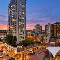 Urban waterfront at dusk with tall illuminated buildings, a marina, and a white tensile canopy in a plaza.