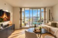 Bright living room with a beige sectional sofa, round coffee table holding a wine bottle and two glasses, and a balcony door showing a city skyline outside.