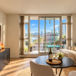 Bright living room with a beige sectional sofa, round coffee table holding a wine bottle and two glasses, and a balcony door showing a city skyline outside.