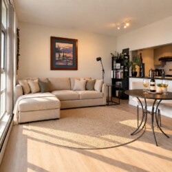 Open-plan living room with a beige sectional sofa, round rug, and a small dining table set for two; kitchen in the background.