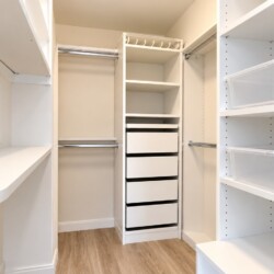 Walk-in white closet with built-in shelves, drawers, and hanging rods, plus clear plastic storage bins on the right side.