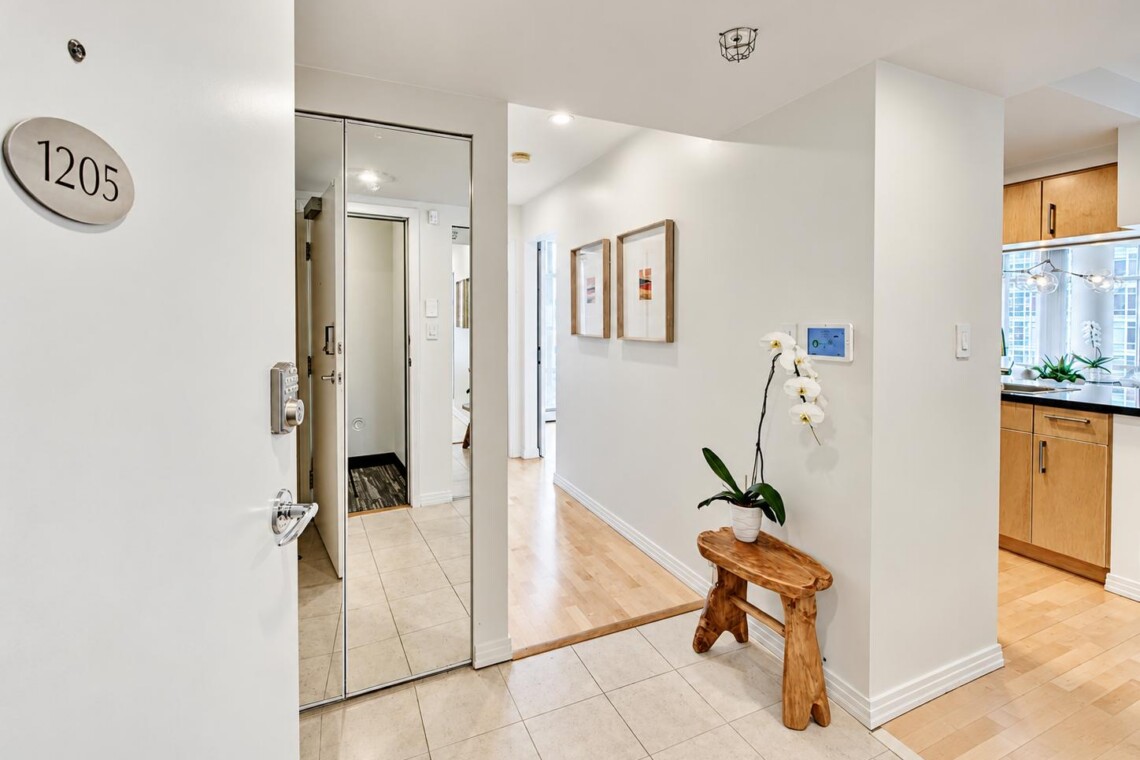 Foyer of an apartment with a 1205 plaque, mirrored wall, wooden bench, and a white orchid plant on a small table edge.